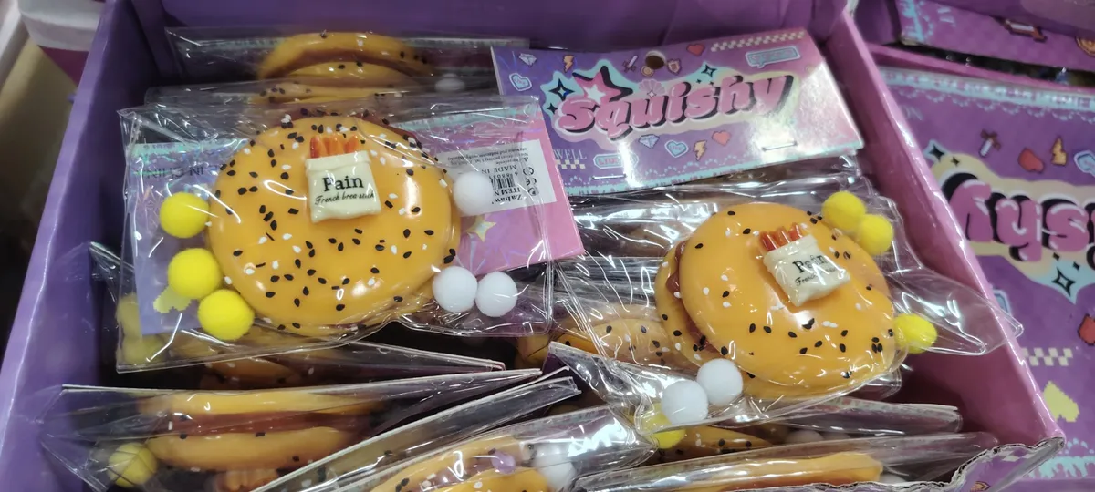 Pomarańczowy squishy hamburger z czarnymi kropkami i pomponami w opakowaniu foliowym