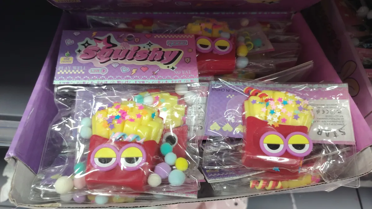 Squishy w kształcie czerwonego kartonika frytek z wielkimi oczami, żółtymi wypustkami i kolorowymi pomponami w przezroczystym