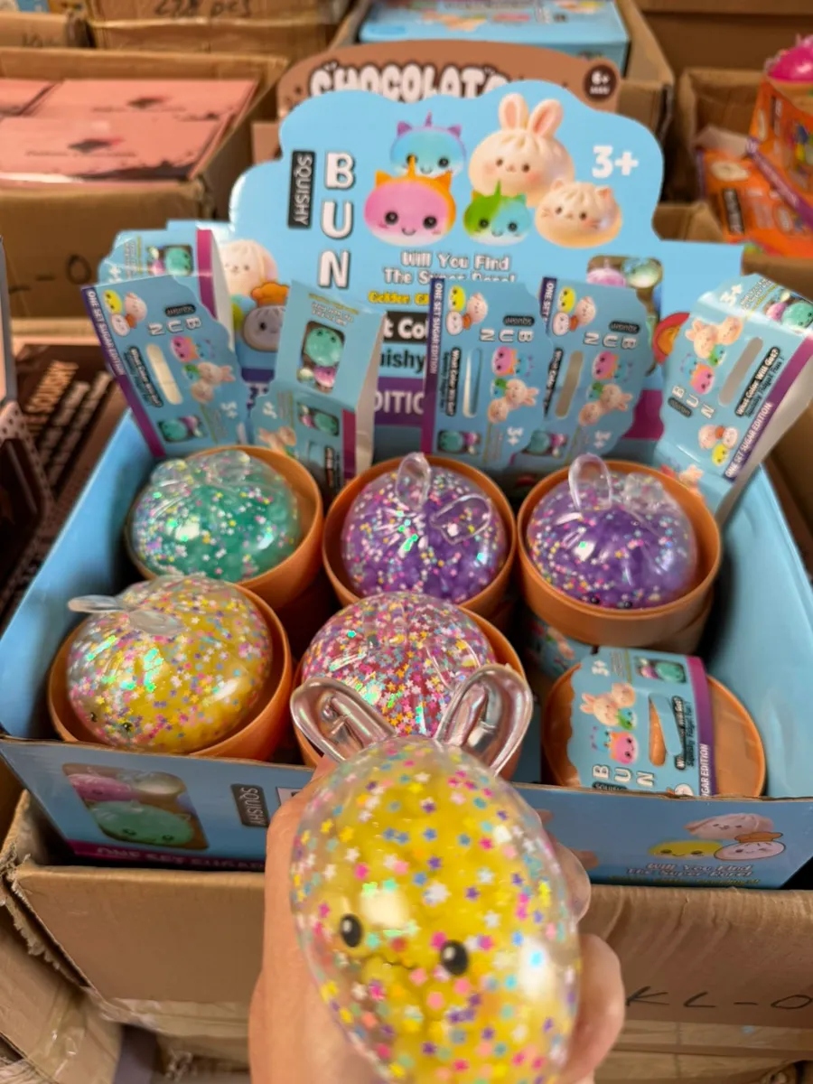 Ręka trzyma żółty squishy Bun z brokatem i konfetti, w tle karton z innymi kolorami serii