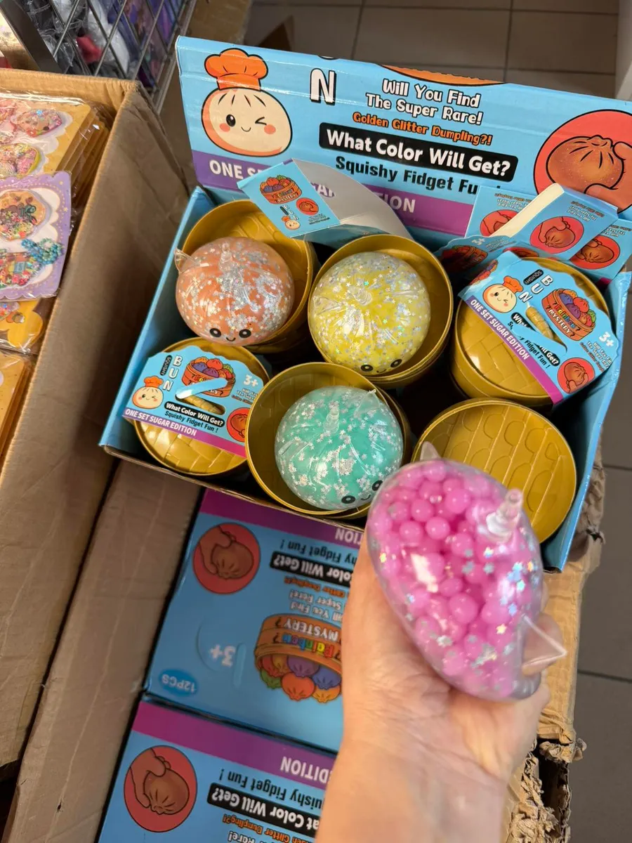Ręka trzyma różowe squishy z brokatem i gwiazdkami, w tle karton z kolorowymi fidget dumpling w złotych pojemnikach