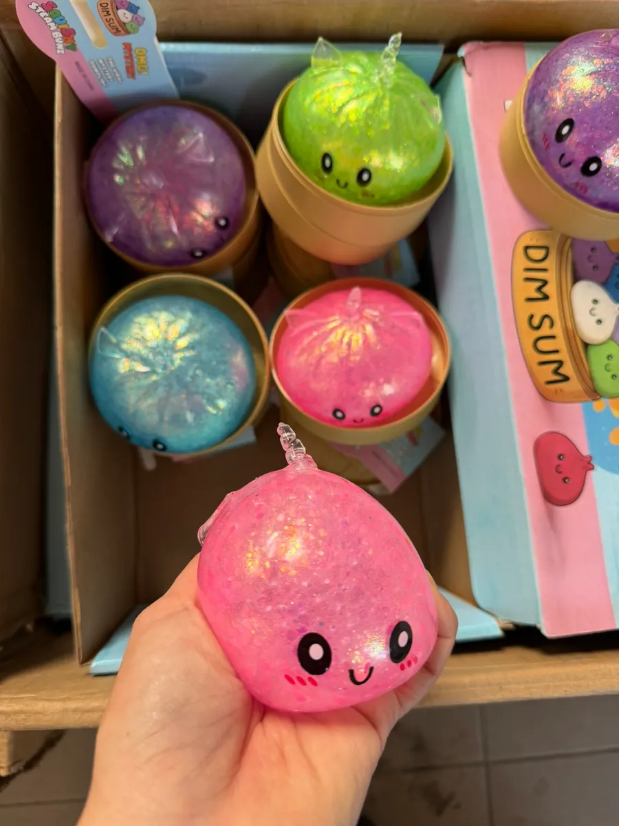 Różowy squishy dim sum z brokatem, czarnymi oczkami i uśmiechem trzymany w dłoni, w tle karton z innymi kolorami