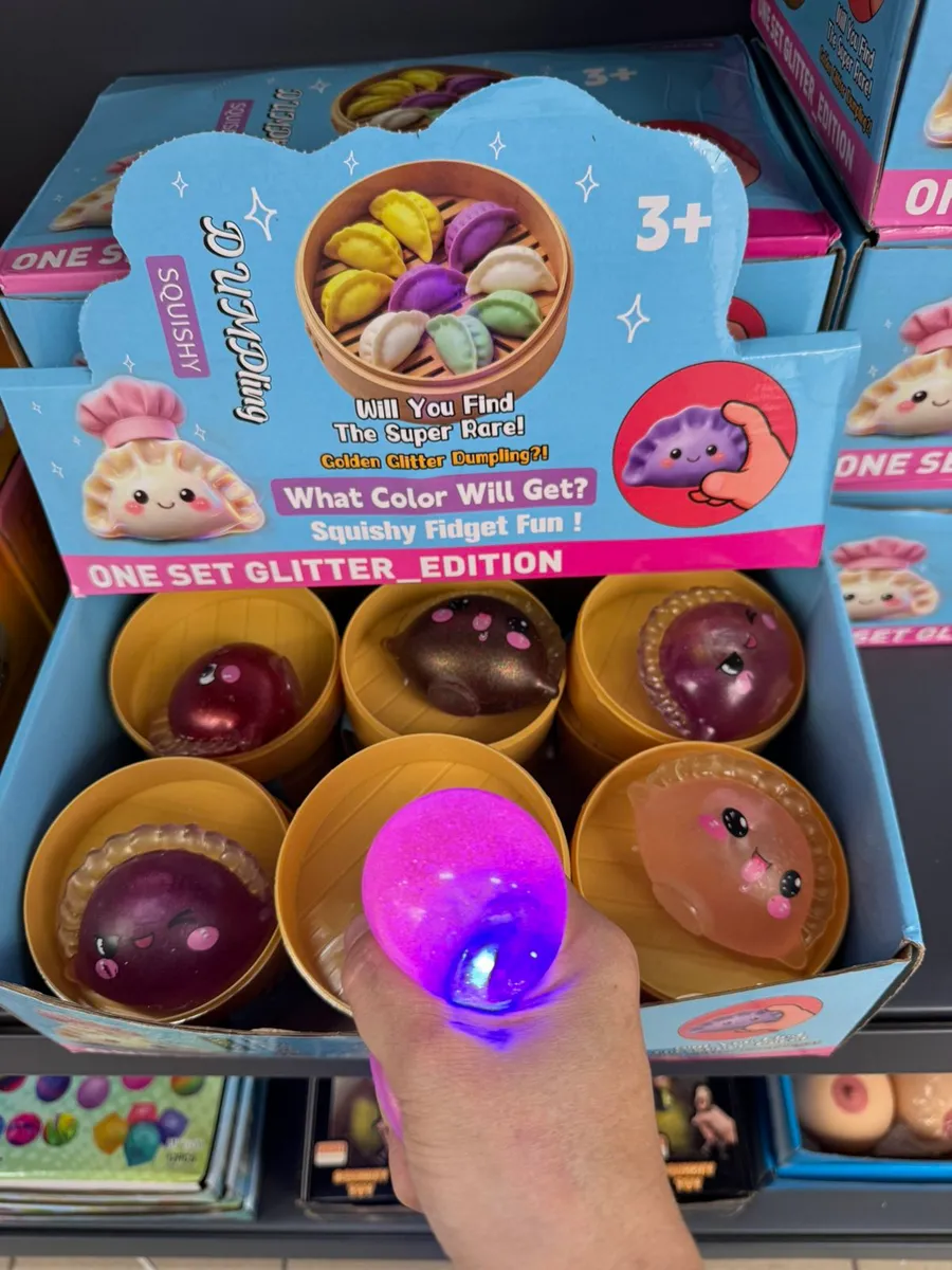 Ręka trzymająca świecący na fioletowo squishy pierożek z brokatem obok kartonowego displaya z innymi kolorami z serii Dumplin