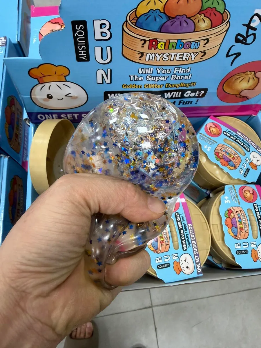Ręka trzyma przezroczysty squishy bun wypełniony brokatowymi gwiazdkami i cekinami na tle kartonów z serii Rainbow Mystery
