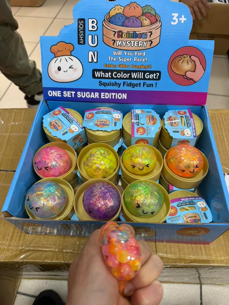 Ręka trzyma kolorowe squishy Bun z brokatem, w tle karton display z wielokolorowymi wersjami Sugar Edition