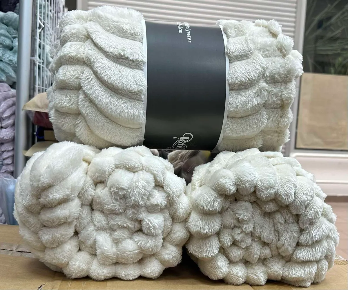 Trzy zrolowane kremowe koce z grubą, falistą fakturą ułożone na kartonie, z czarną banderolą z napisem Polyester i logo Daro