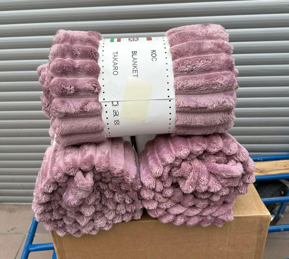 Złożony w kostkę puszysty koc w pudrowym różu z szerokimi pasami, zapakowany w papierową etykietę z napisami KOC, BLANKET, TA