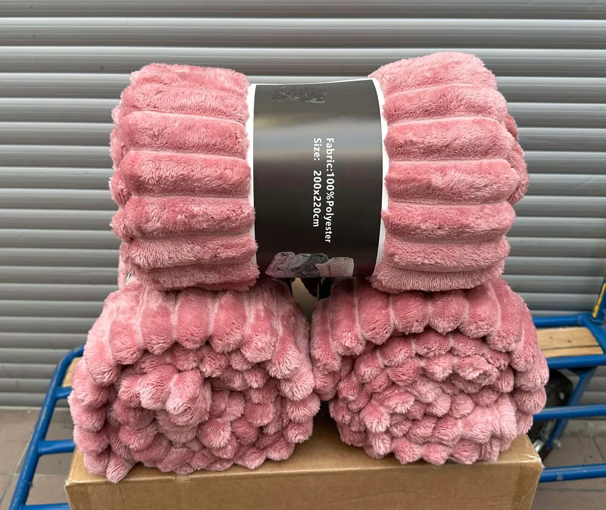 Zrolowane pudroworóżowe koce z puszystą fakturą dużych pasów, opakowane w czarną etykietę z napisem Fabric 100% Polyester Siz