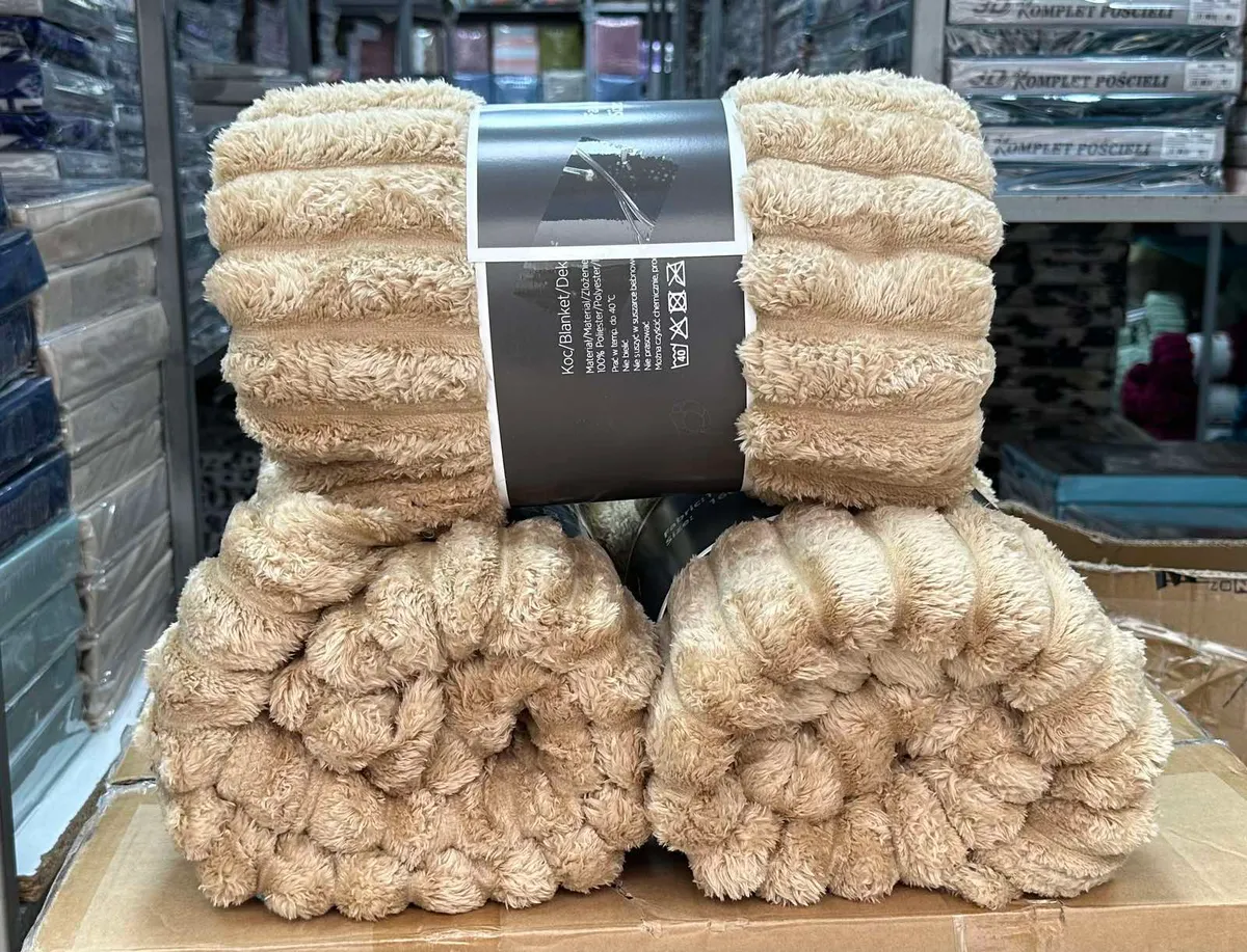 Złożony beżowy koc z grubymi pasami i etykietą produktową, widoczna puszysta faktura materiału