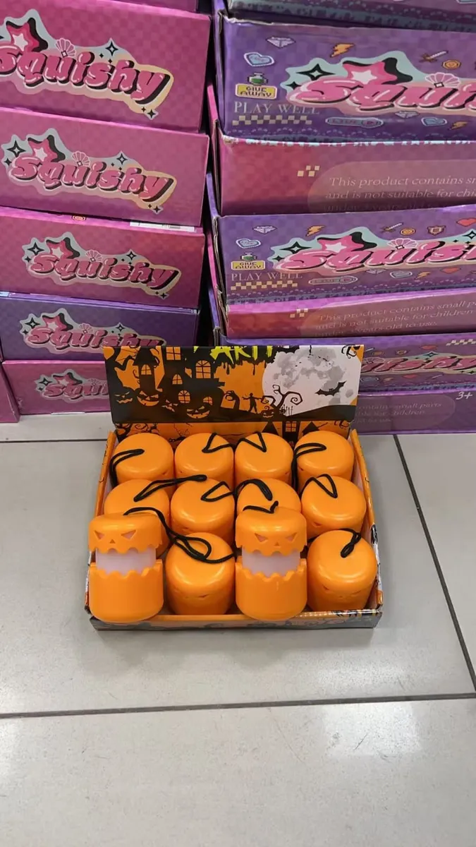 Pomarańczowe lampiony dynie z czarnym sznurkiem w kartonie na tle różowo-fioletowych opakowań Squishy
